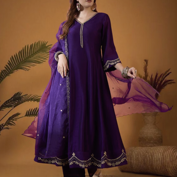 "Woman wearing a royal purple embroidered kurta set with matching dupatta, accessorized with traditional silver jewelry, sitting on a wooden chair against a beige background."